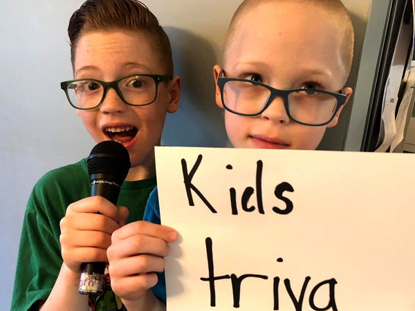 Two boys are in a picture. The one of the left holds a mic to his mouth, the one on the left holds up a sign that says "Kids trivia."