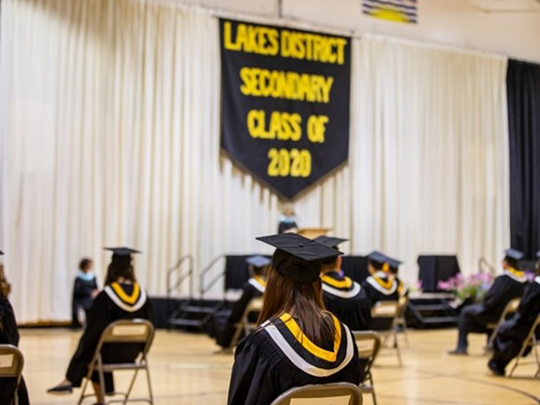 Students in gowns sit a safe distance apart, listening to graduation speeches.