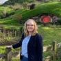 Woman with shoulder length blonde hair and glasses stands in front of hobbit house