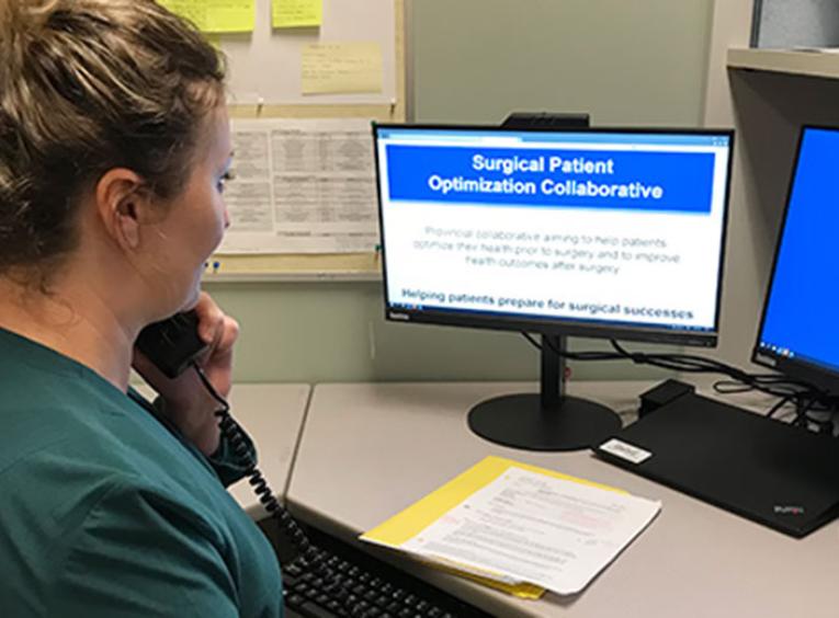 A side angle shot of a woman in a green sweater sitting at a desk in front of a computer screen that says "Surgical Patient Optimization Collaborative."
