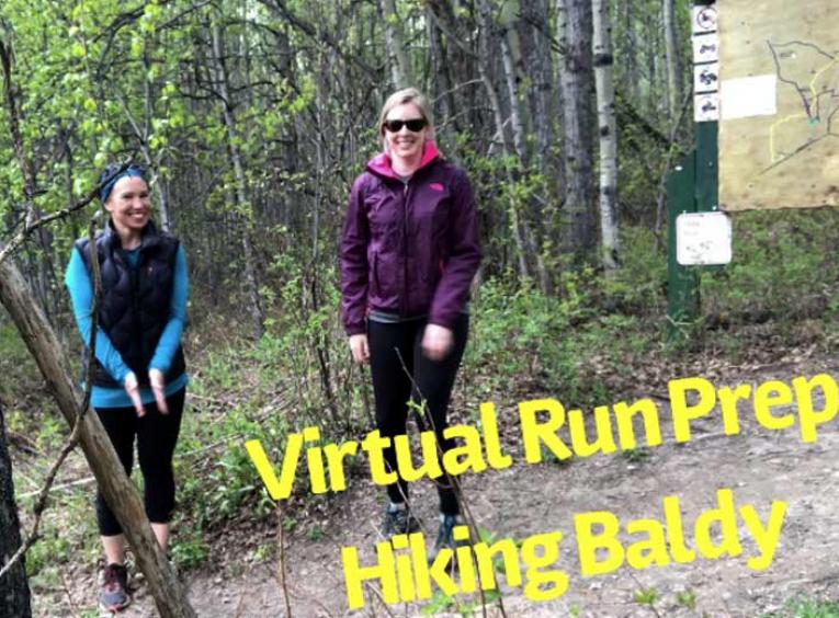 Two women in hiking apparel smile into the camera as they hike a wooded trail.