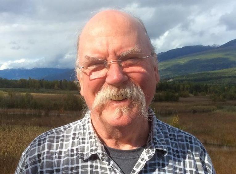 Elderly man with glasses and a large moustache stands in field with mountains in the background