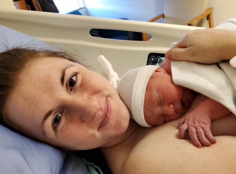 A smiling woman lays in a hospital bed with a newborn against her chest.