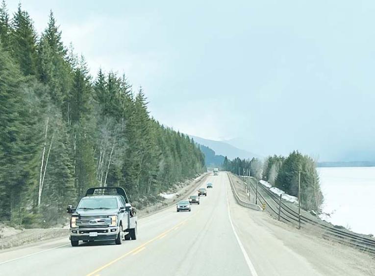 a highway with a mountain on one side and a lake on the other. A stream of traffic is in the left-hand lane. 