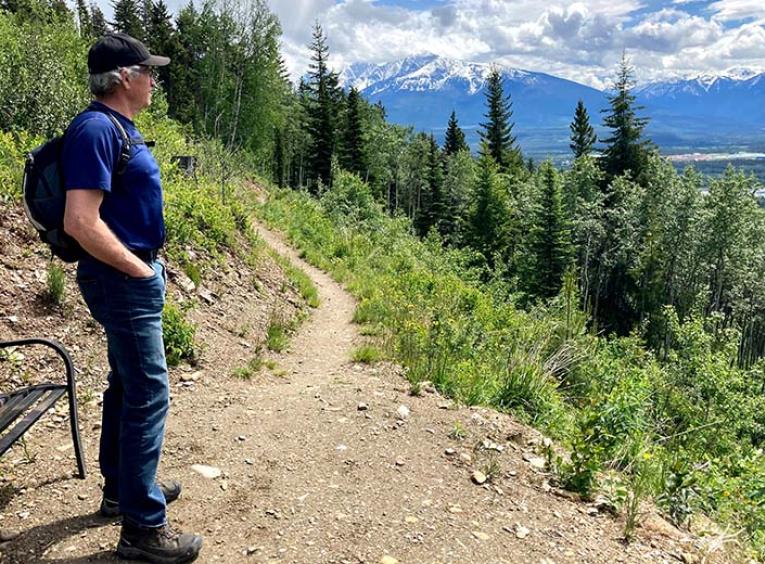 a man in a blue shirt stands on the edge of a cut bank looking over the tree lined landscape